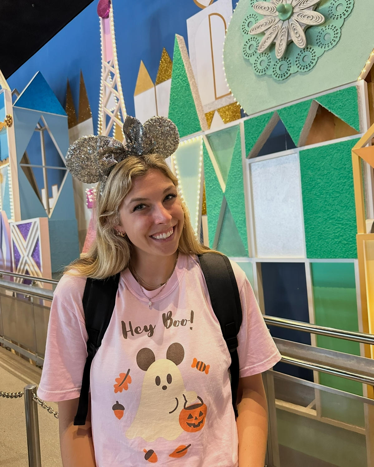 A woman wearing a pink Mickey Mouse Halloween t-shirt and gray Mickey ears stands smiling in front of a colorful Disney-themed mural.