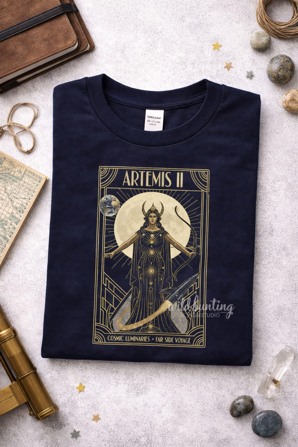 Navy blue t-shirt with gold graphic design featuring a Artemis ii figure and text, placed on a textured surface with decorative items.