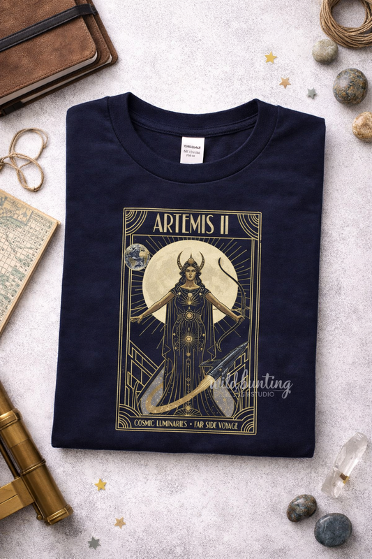 Navy blue t-shirt with gold graphic design featuring a Artemis ii figure and text, placed on a textured surface with decorative items.