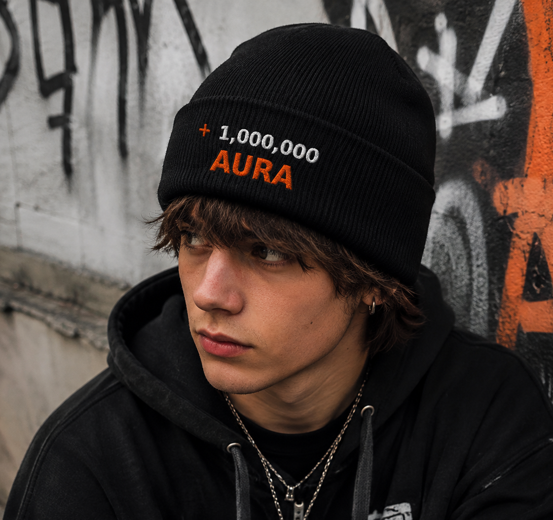 A young man wearing a black knit beanie with '1,000,000 AURA' embroidered on it, paired with a black graphic hoodie, standing against a graffiti-covered wall.