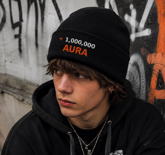 A young man wearing a black knit beanie with '1,000,000 AURA' embroidered on it, paired with a black graphic hoodie, standing against a graffiti-covered wall.