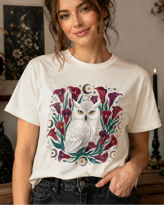 A woman wearing a cream-colored graphic t-shirt with a detailed illustration of an owl surrounded by purple calla lilies and green leaves, set against a cozy, styled room background with candles and dried flowers.