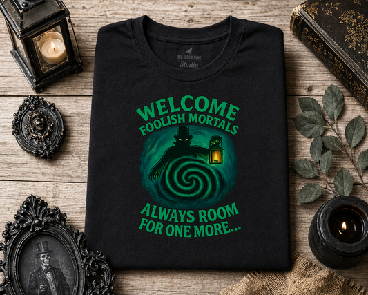Black t-shirt with green graphic text that says Welcome Foolish Mortals and text on a wooden surface with decorative items.