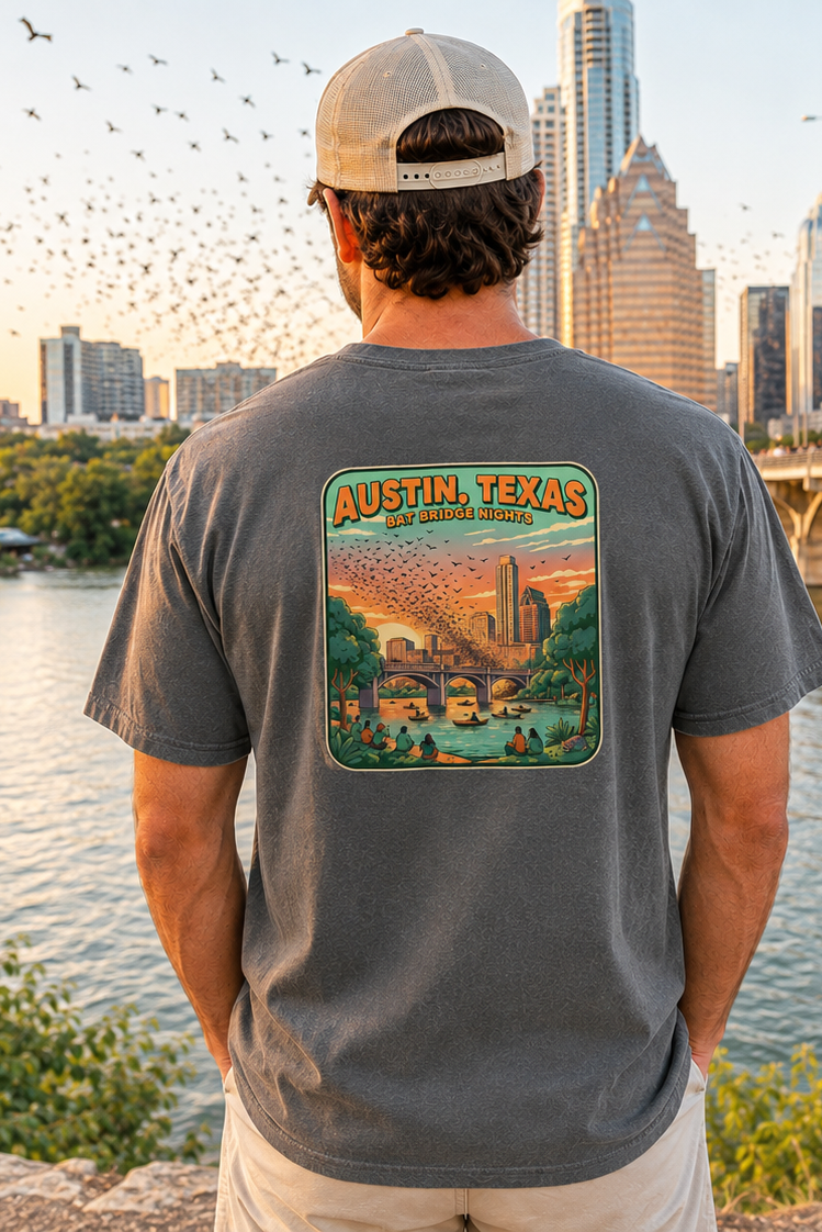 A man stands with his back to the camera wearing a dark gray t-shirt featuring a colorful 'Austin, Texas' graphic on the back, set against a scenic city skyline and river at sunset.