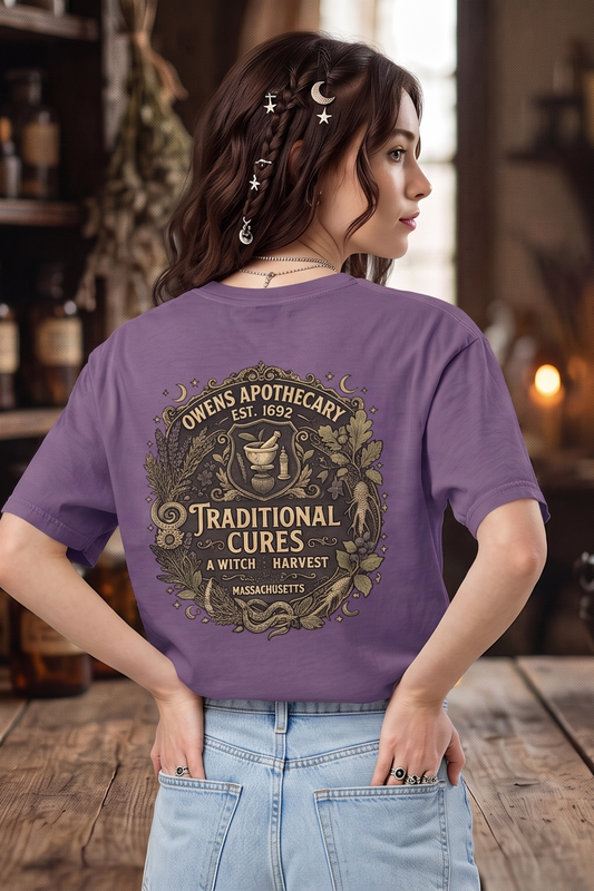 A woman stands with her back to the camera in a rustic apothecary shop, wearing a purple graphic t-shirt with a circular vintage-style design that reads 'Omens Apothecary 2023 Traditional Cures Witch Harvest' and light blue jeans.
