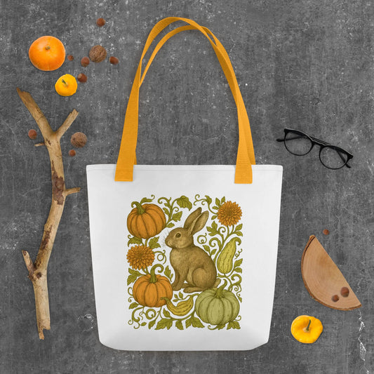 White tote bag with orange handles featuring a vintage-style rabbit surrounded by pumpkins, gourds, and autumn flowers, displayed on a rustic gray background with fall props.